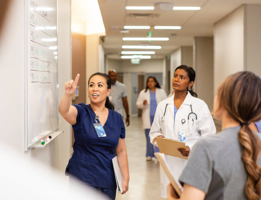 Young adult female nurse gestures towards the whiteboard while meeting with her team